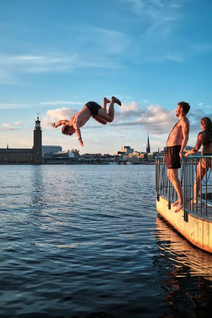 En person gör en volt ut i vattnet med Stadshuset i bakgrunden medan två andra tittar på från en brygga